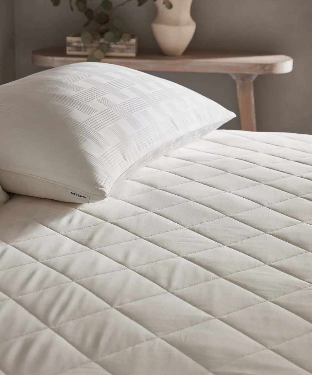 White Cozy Earth Bamboo Mattress Pad shown on a mattress with a white pillow on top. Behind is a small wooden side table with a vase and houseplant, against a grey wall.