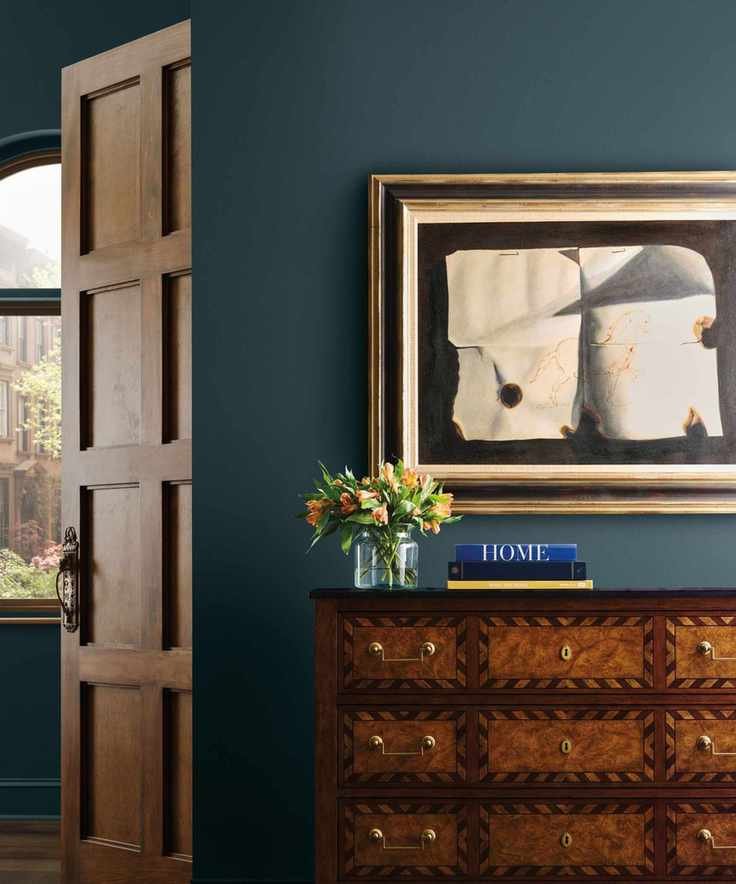 Dark navy painted walls with chest of drawers, abstract art on wall, stack of books and vase of flowers