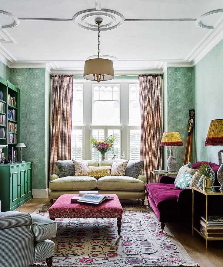 Living room with wooden floor and green walls, purple and green sofas with cushions, central ottoman on a rug below ceiling with coving.