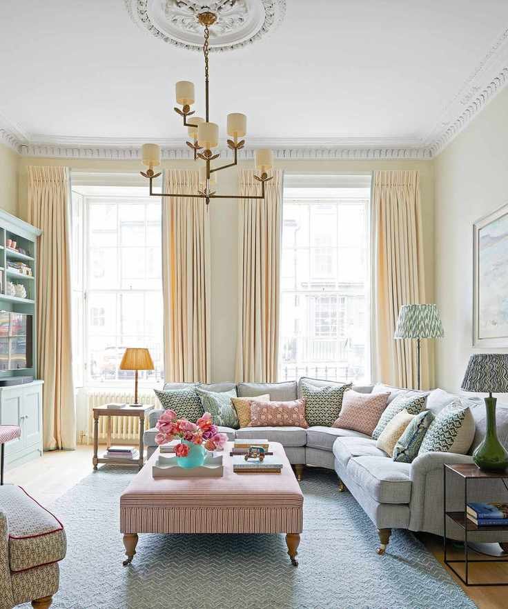 Contemporary living room with large floor to ceiling windows, cream drapes, white painted ceiling cream painted walls, gray corner sofa, wooden floors, blue rug, large ottoman with wooden feet, metal chandelier with lamp shades