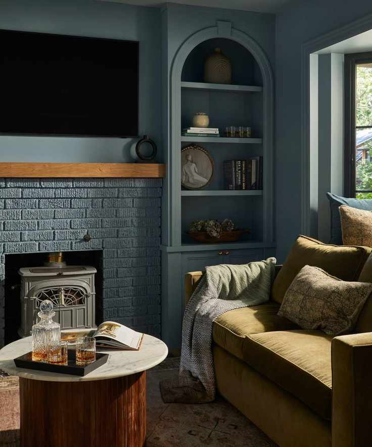 a blue color drenched living room with arched niche storage, a blue painted brick fireplace, green couch and a round marble topped coffee table