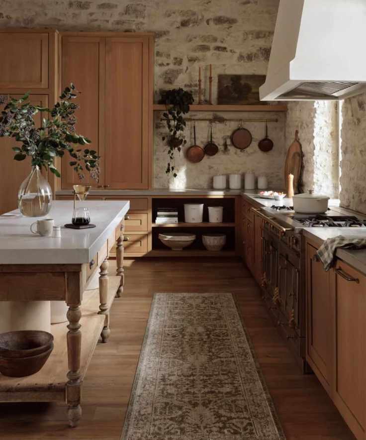 farmhouse kitchen with exposed stone walls, oak cabinetry, a plaster hood, wooden work table island with marble countertop top and a vintage style runner rug