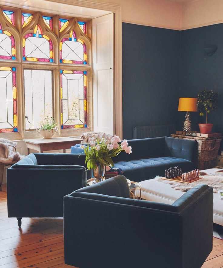 Living room with dark blue walls, wooden floor and dark blue velvet armchairs and sofa, leaded stone window with stained glass in the background.