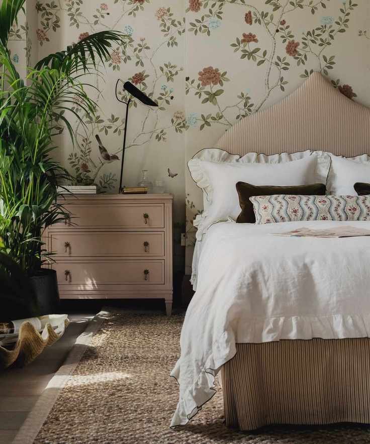 floral romantic bedroom with floral wallpaper, a pink upholstered bed, white ruffled scalloped bedding with a floral bolster cushion