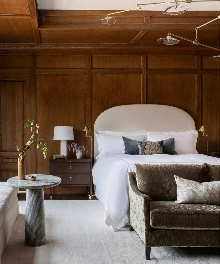 calm timeless bedroom with original dark wood panelling, a large neutral rug, neutral bed, dark green velvet couch at the foot of the bed and a small marble table