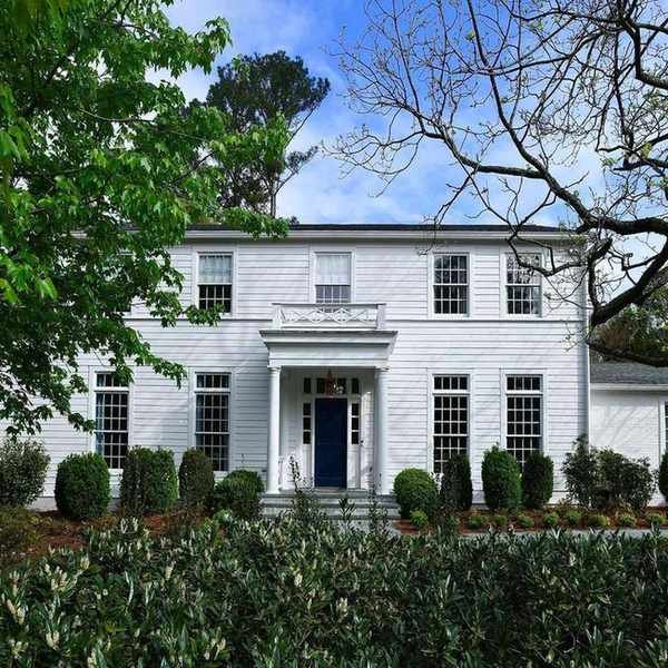 A large traditional home with a white exterior and Georgian-style windows. A dark blue front door with a porch with pillars and lots of greenery in the foreground.
