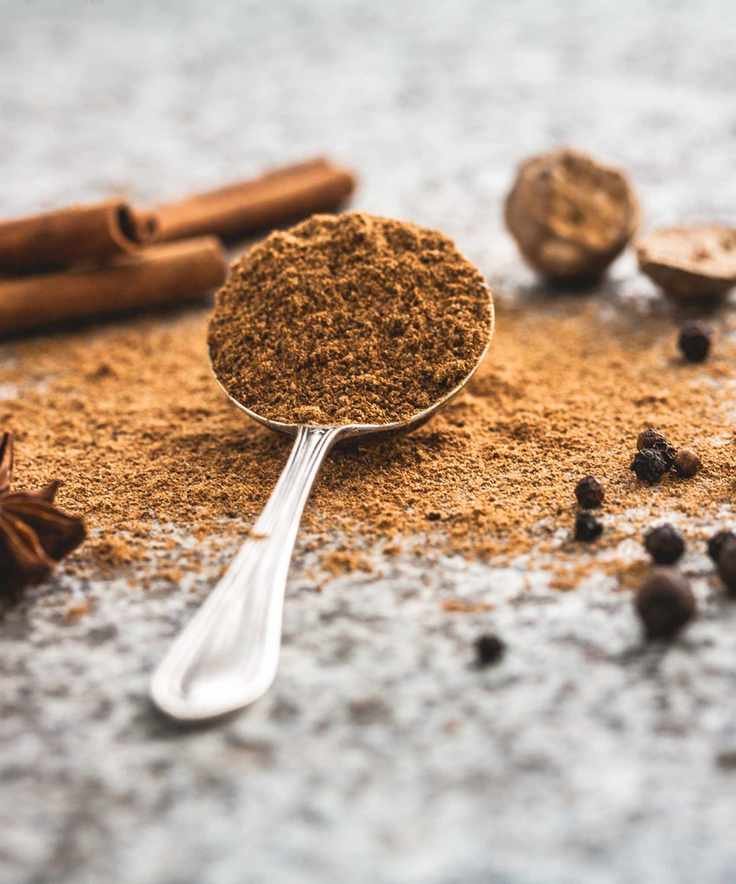 Silver spoon of cinnamon powder, on a granite surface, with loose cinnamon powder and sticks in view