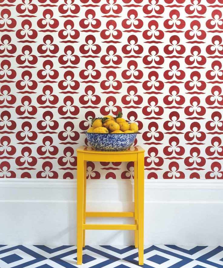 A blue bowl filled with lemons on a yellow stool. Behind is a red and white patterned wallpapered wall, oversized baseboard, and blue and white patterned floor.