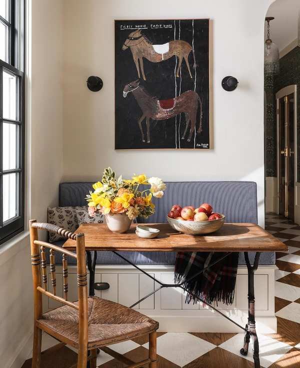 A photo of a window seat with a built-in banquette below a folksy painting of horses.