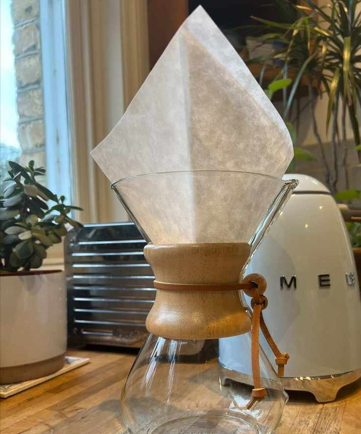 A glass CHEMEX coffee maker with a white filter. In the background is a small white pot with a small plant, silver Dualit toaster, baby blue Smeg kettle, and large houseplant with a baby blue backsplash and wooden corner cabinet. To the left is a window, surrounded by yellow brick on the outside.