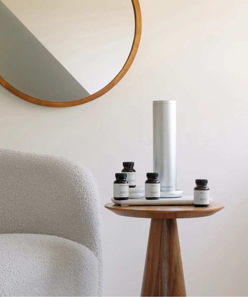 White AromaTech AroMini on a wooden table with four essential oil bottles with a boucle chair and circular mirror against a white wall.