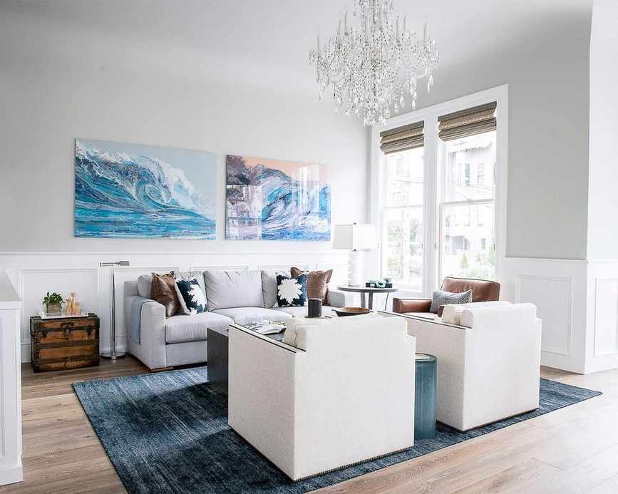 Coastal living room with two pieces of artwork on wall, light gray painted wall, large glass chandelier, blue rug over wooden floor