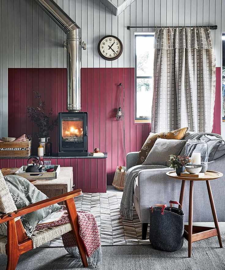 Large living room with wooden paneled walls, painted in gray and dark red paint, raised level with wood burner, gray carpet, gray patterned rug, sofa with cushions and throws, wooden mid-century lounge chair, wooden coffee table, windows with patterned curtains, round clock on wall