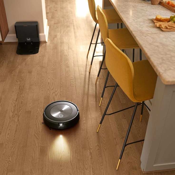 iRobot Roomba j7+ robot vacuuming on a wooden floor in a kitchen, with the robot's cleaning base visible in the background.