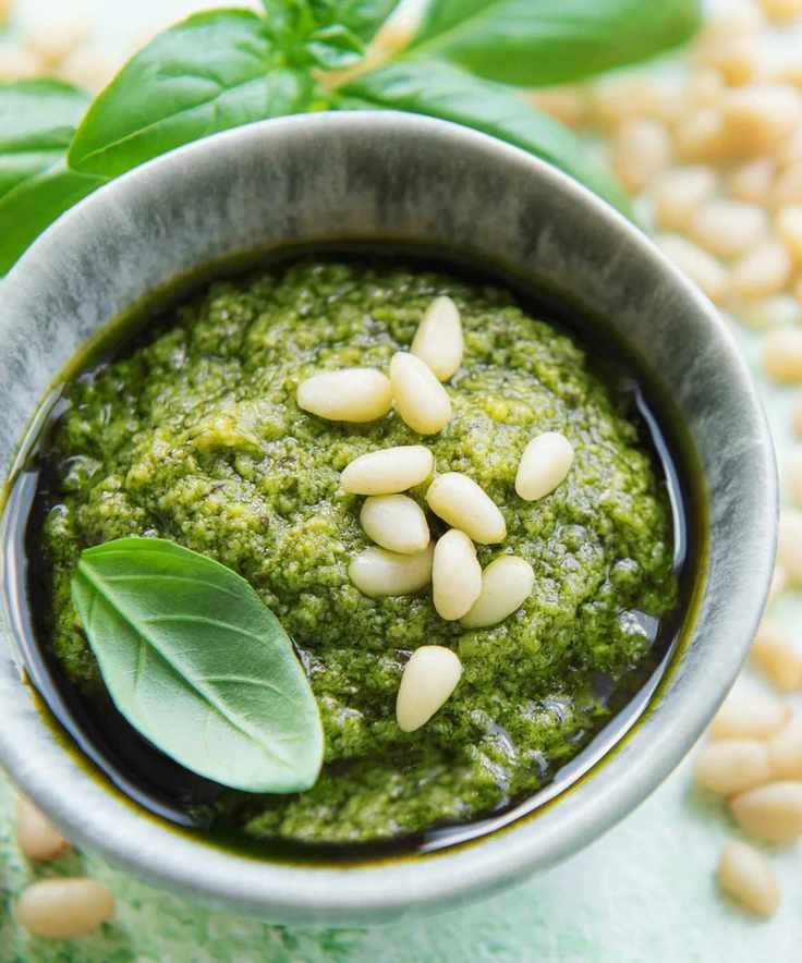 A pot of fresh green pesto with pine nuts and garlic scattered nearby