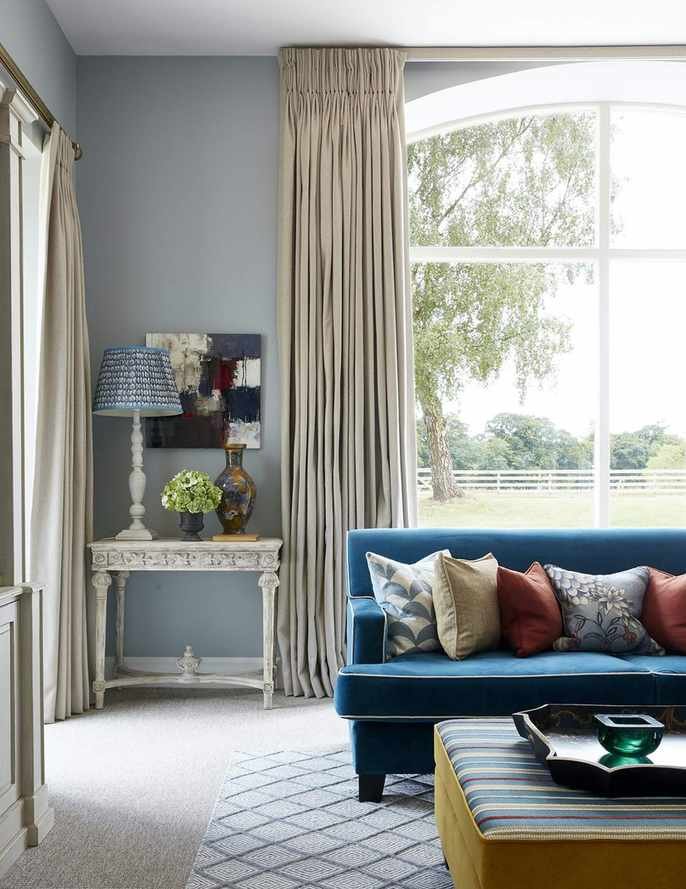 An example of small living room decor ideas showing a blue sofa in a sitting room with blue walls and cream coloured drapes.