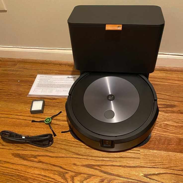 The iRobot Roomba j7+ robot vacuum, cleaning base, brush, cable, manual, and filter on wooden floor