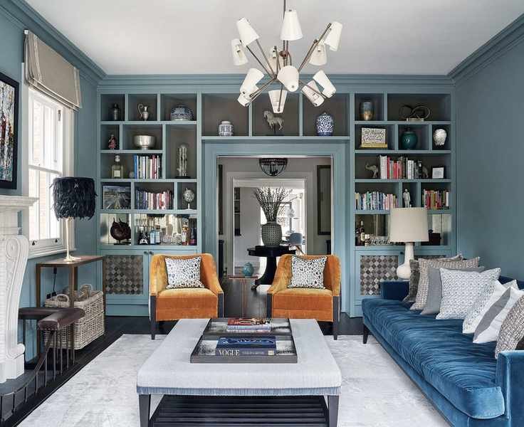 Spacious blue-painted living room featuring floor-to-ceiling storage and display units, two orange chairs, blue sofa with gray cushions, large gray rug, coffee table with ottoman, fireplace with art above, and modern chandelier