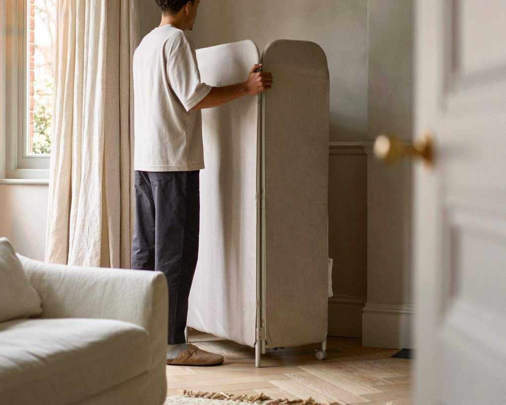 A man next to the Eclipse&amp;trade; 3-tier Ecru Clothes Drying Rack With Screen in a bright living room