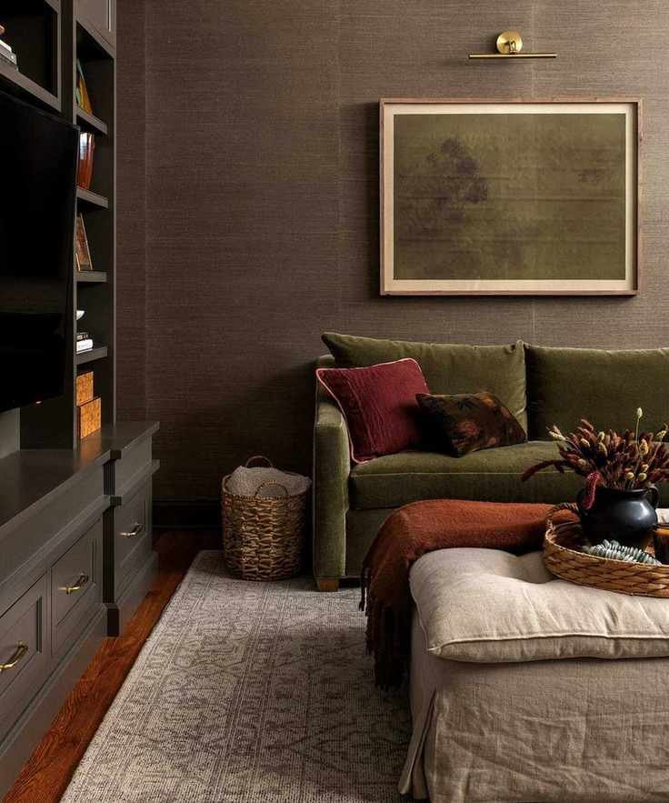 Dark living room with patterned rug, green sofa with cushions, framed minimalist painting, partial black bookshelf to the left, footstool, wooden floors.