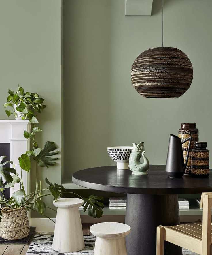A matte green dining room with a round black dining table, wooden chairs, and a low hanging black pendant light