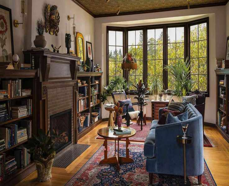 a large home library with original tudor bay window, a large vintage rug, blue sofa, accent wallpapered ceiling and wooden fireplace with bookcase built-ins