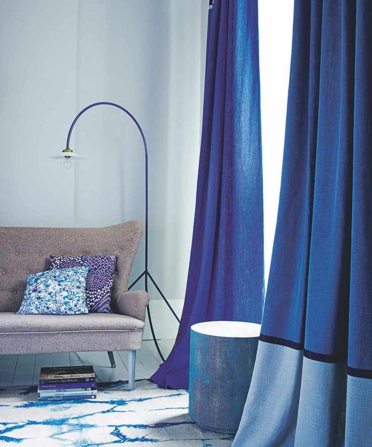A corner of a pale blue room with blue curtains, a blue and gold stool, brown couch with patterned cushions, a blue curved floor lamp, white and blue patterned rug, and stack of coffee table books.