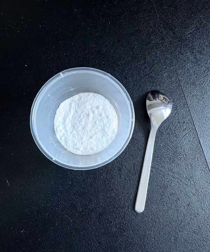 A round plastic container with baking soda inside. A silver teaspoon beside it.