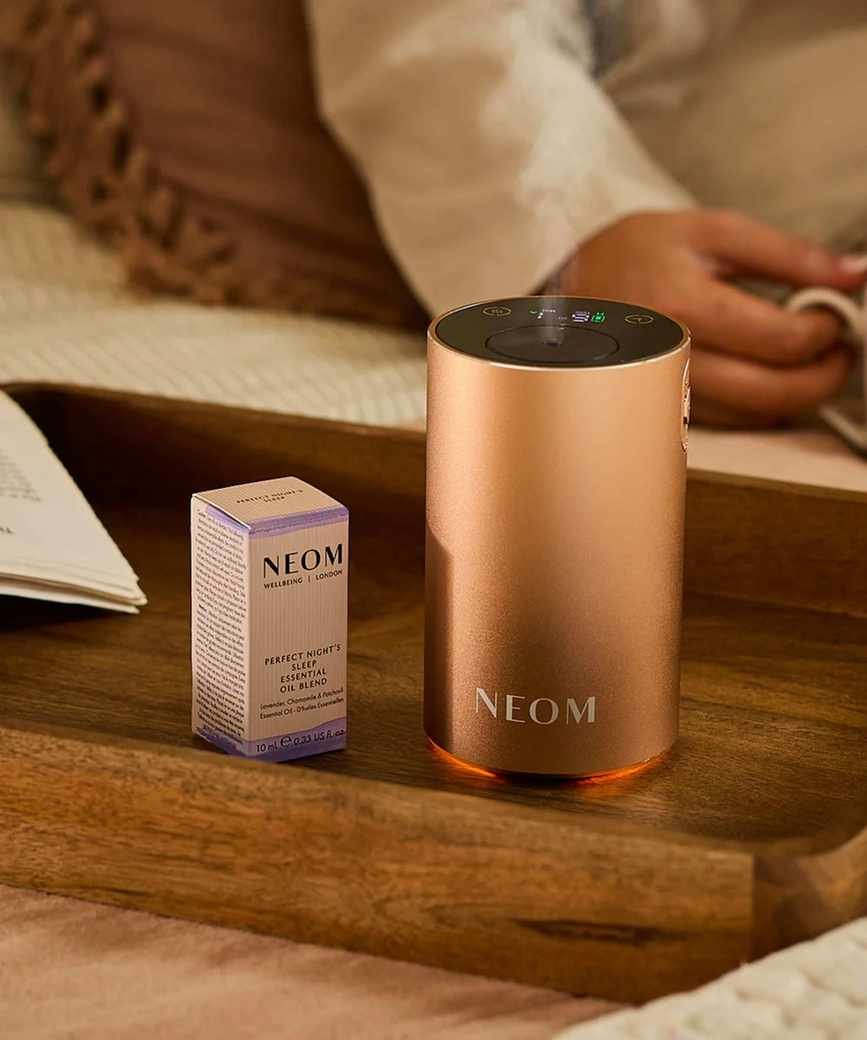 A rose gold Neom Wellbeing Pod Mini+ on a wooden tray, beside a pale pink and purple cardboard packaging box of essential oil.