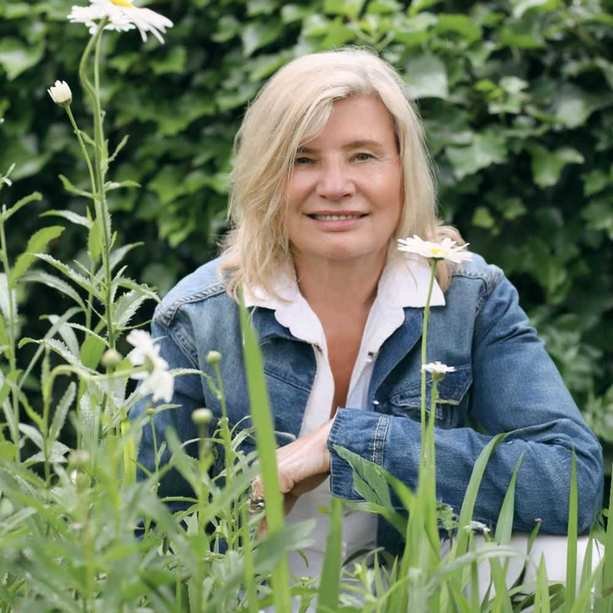 A headshot of Alicia Sokowlowski wearing a white shirt and blue denim jacket sitting behind grass and daisies with a green leafy background