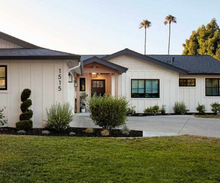 A bungalow home exterior with warm white siding, a dark brown door, and a light brown porch. Dark window trim and a gray roof with a grass lawn and paved driveway.