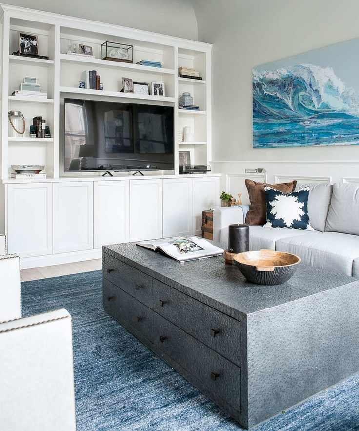 Bright white living room showcasing a white storage unit with a TV, a large gray coffee table with drawers, a blue rug, and wall art featuring a wave design