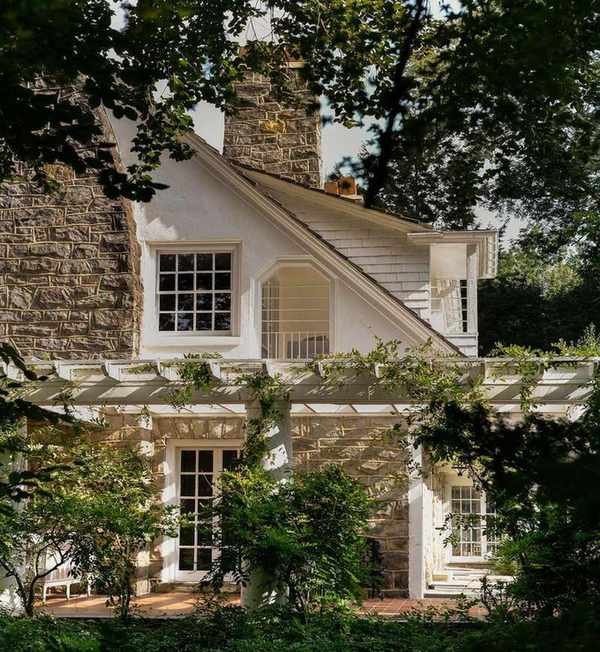 A traditional home exterior with exposed stone, white paint, and traditional-style windows, surrounded by greenery from trees.