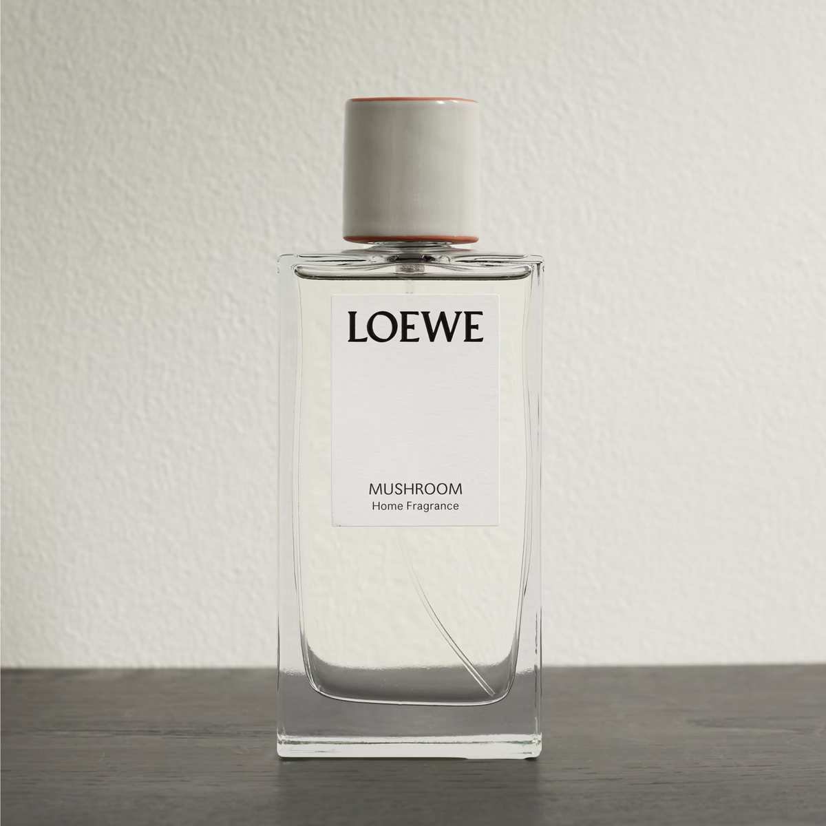 A glass bottle of Loewe Mushroom room spray against a white wall on a wooden surface