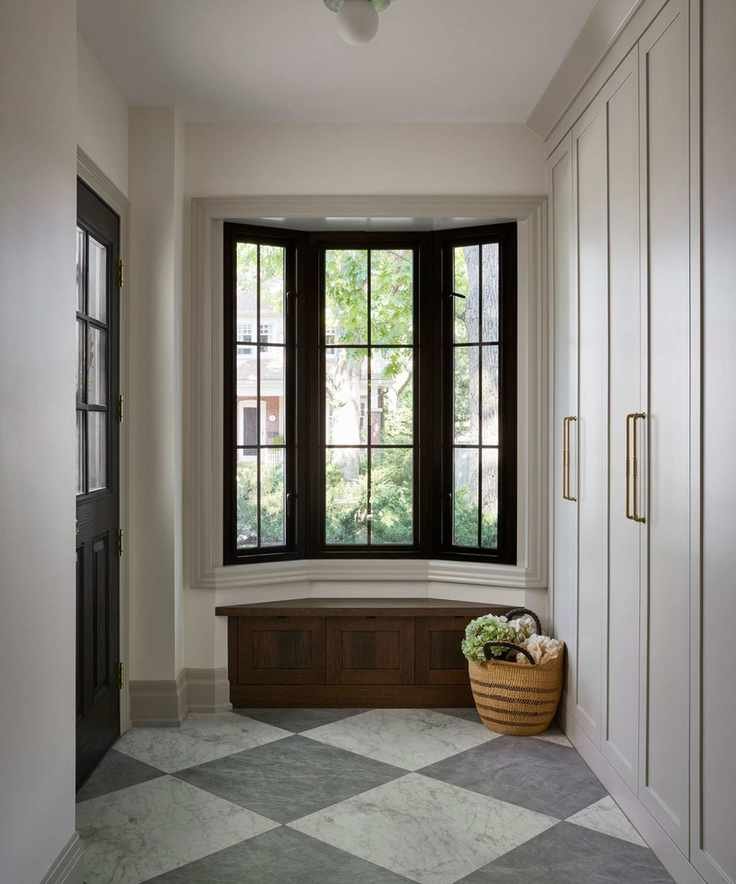 a bright entryway with a large bay window and dark wooden window seat, grey and white marble checkerboard floor, and custom mudroom joinery
