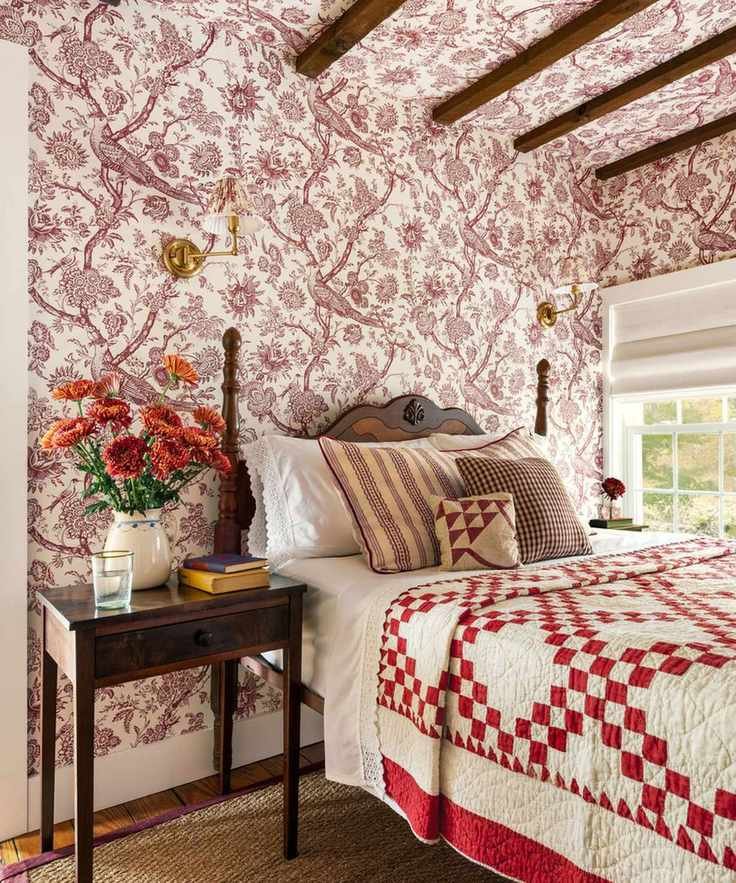 A cozy, traditional bedroom featuring burgundy floral toile wallpaper that extends onto the ceiling. The bed is dressed with a red and white checkered quilt and mixed pattern pillows, flanked by a dark wood nightstand with orange flowers and brass wall sconces