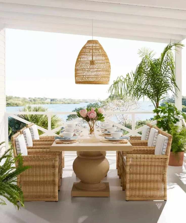 coastal garden veranda with a sea view by serena and lily with a large pedestal wooden table with bamboo and wicker armchairs and a seagrass ceiling pendant