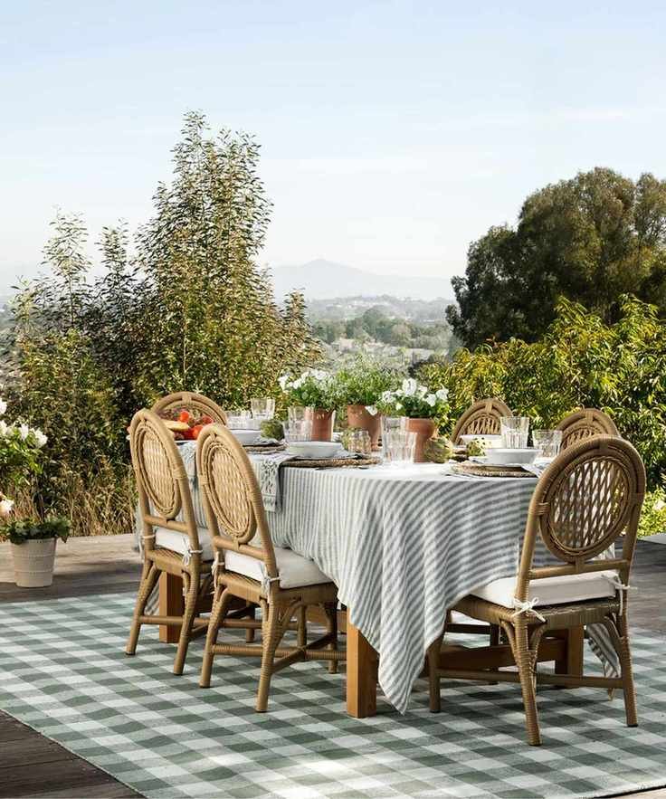 outdoor dining table with wooden and rattan furniture, a green striped linen tablecloth and a green plaid rug surrounded by potted plants