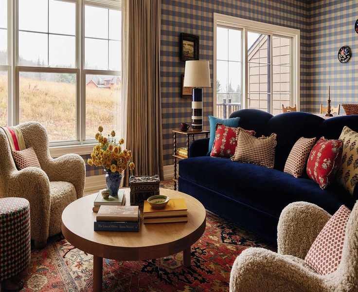 an open plan living area in a home in the Catskills with blue gingham wallpaper, a navy velvet couch, vintage red rug, sheepskin armchairs and a round wooden coffee table