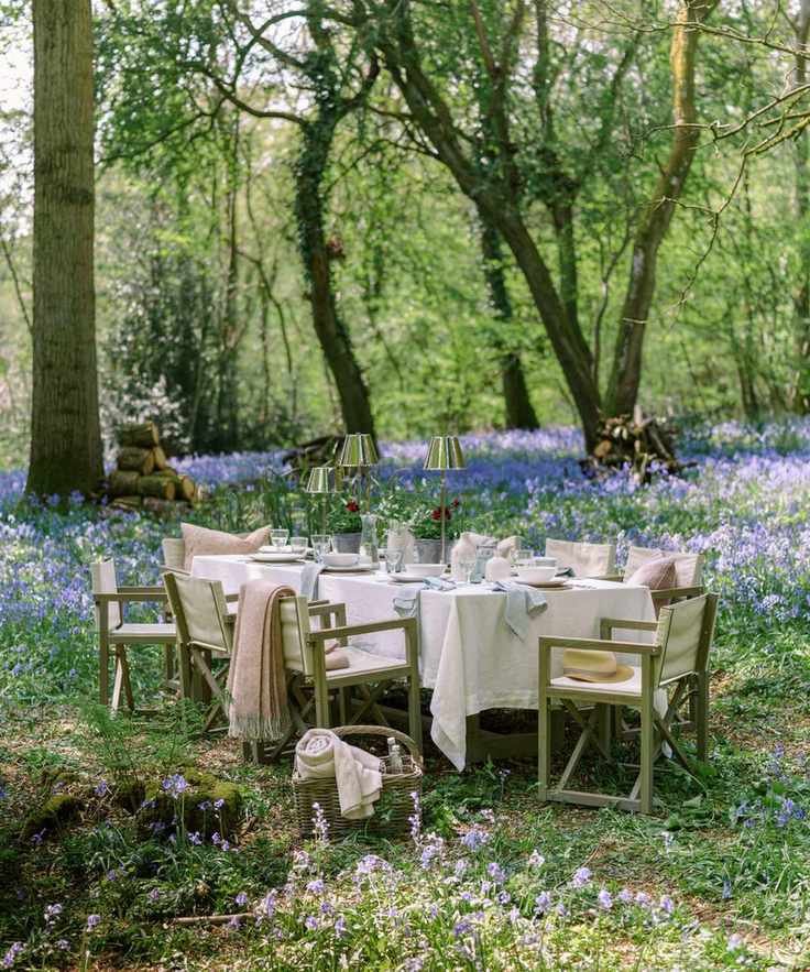 large outdoor table set up in a woodland with bluebells the table is laid with white linens and silver portable table lamps