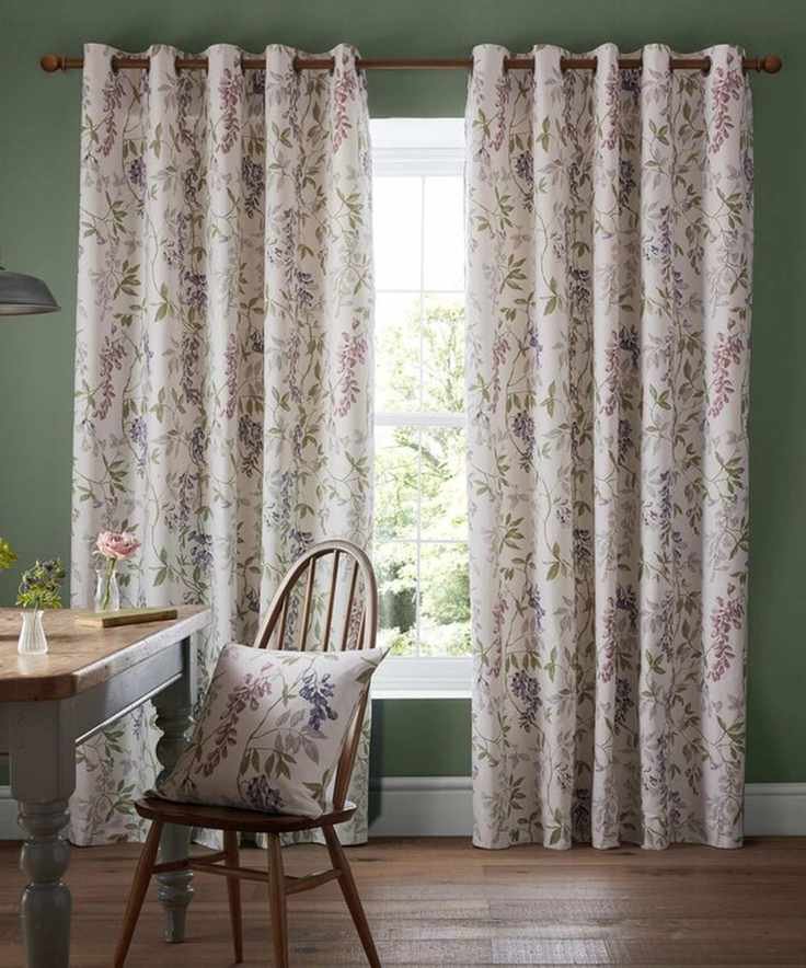 Floral patterend curtains hanging either side of late white framed window, set into a green painted wall. A wooden country-style dining chair with a matching pillow on it.