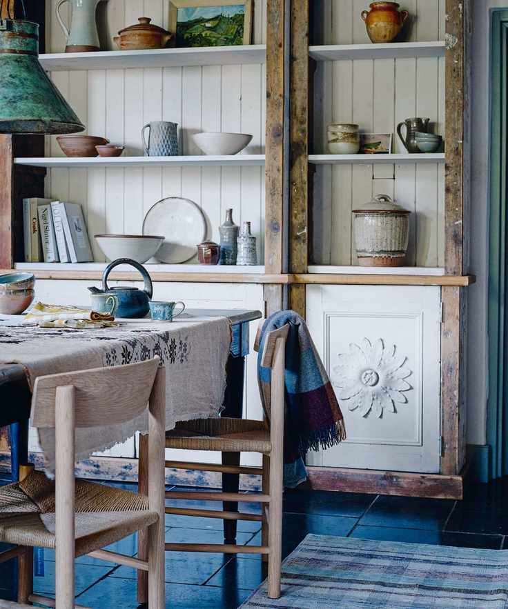 Kitchen table with an embroidered tablecloth and a dresser in the background displaying china dishes and jugs.