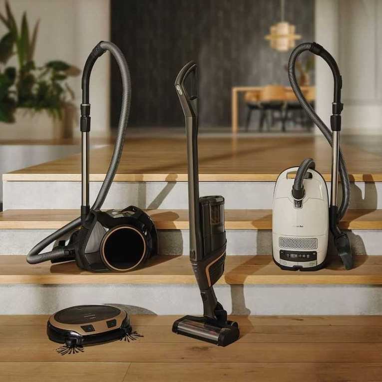 Four Miele vacuum cleaners – a cordless vacuum, robot vacuum and two canister vacuums – on set of wooden stairs in luxury home, with plants and furniture pictured in background.